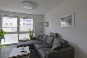 A modern living room with a grey leather sofa, a coffee table with a remote control, and a large window with blinds. - Holiday home in Cuxhaven with swimming pool (Cuxhaven)