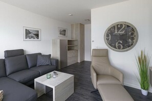 A modern living room with a grey sofa, a beige armchair, a coffee table, and a large wall clock. - Holiday home in Cuxhaven with swimming pool (Cuxhaven)