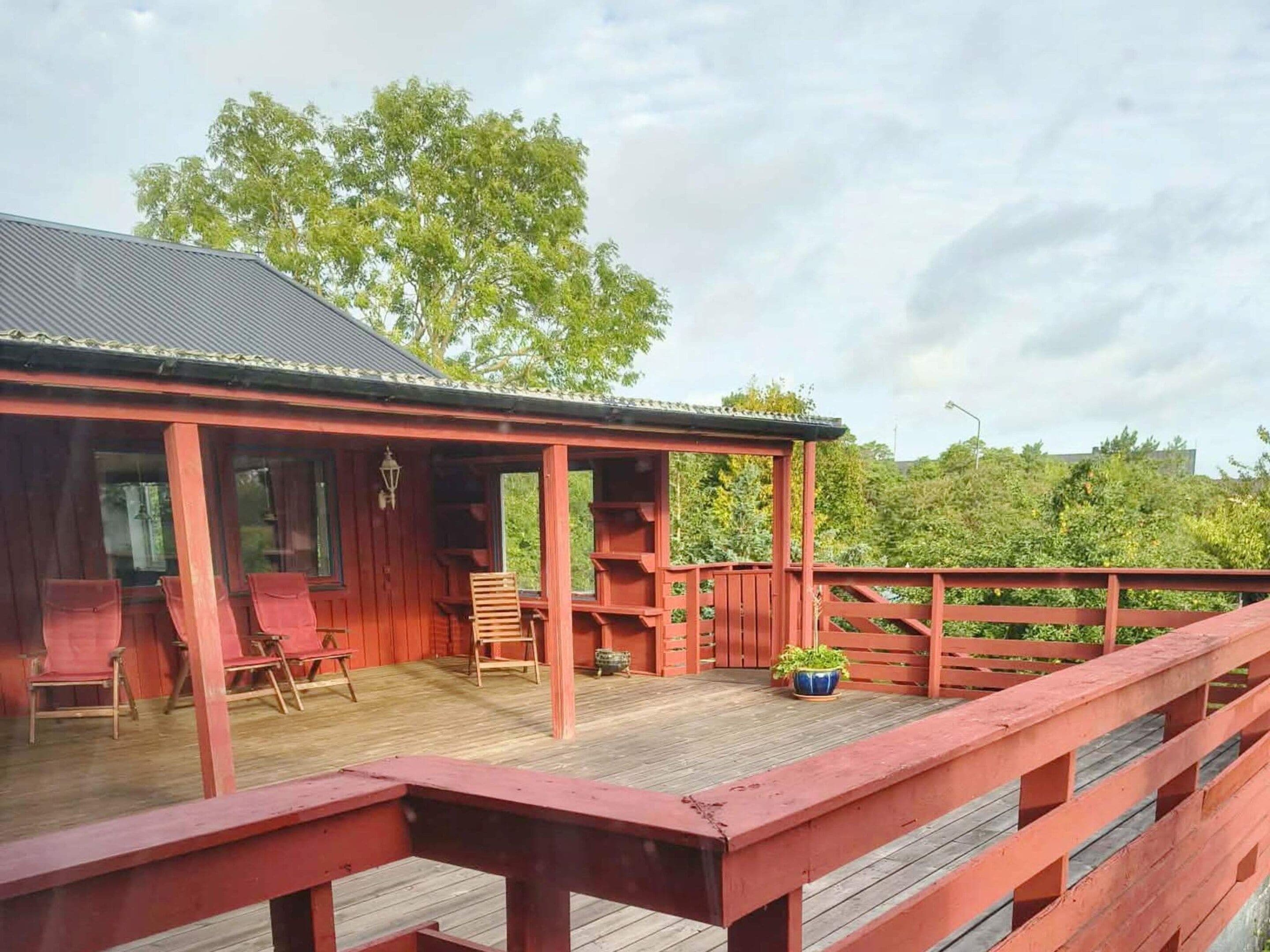 A wooden deck with red railings, a covered seating area with chairs, and a potted plant.