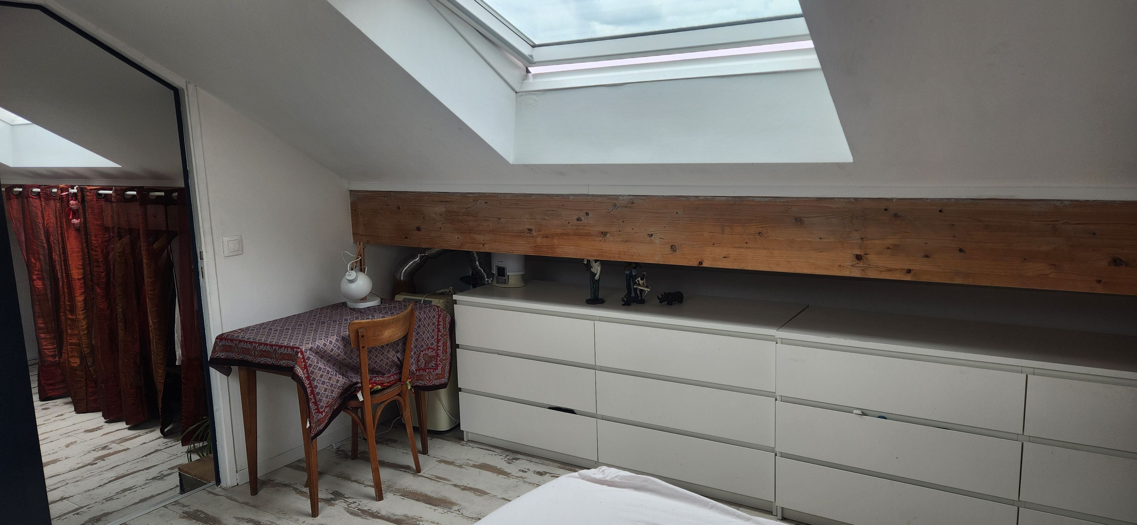 Quarto pequeno e compacto com teto inclinado e claraboia, com uma mesa, cadeira e um armário branco.
