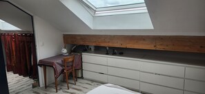 Ein kleiner, kompakter Raum mit schrägem Dach und Dachfenster, ausgestattet mit Schreibtisch, Stuhl und einem weißen Schrank.