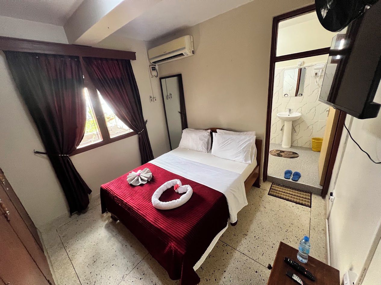 A hotel room with a bed, a red bedspread, a white heart-shaped decoration, a window with curtains, an air conditioning unit, a mirror, and a sink with a yellow bucket.