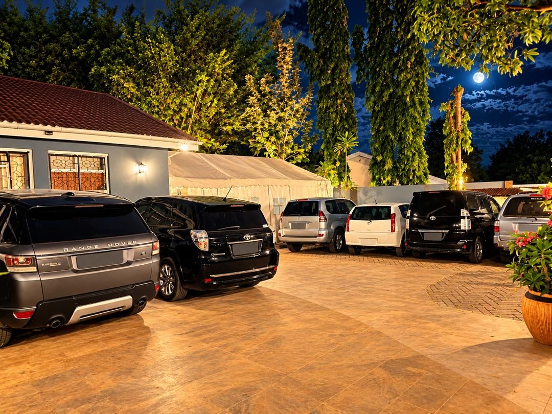 A parking area with multiple cars, a building with a tiled roof, and a large tree in the background.
