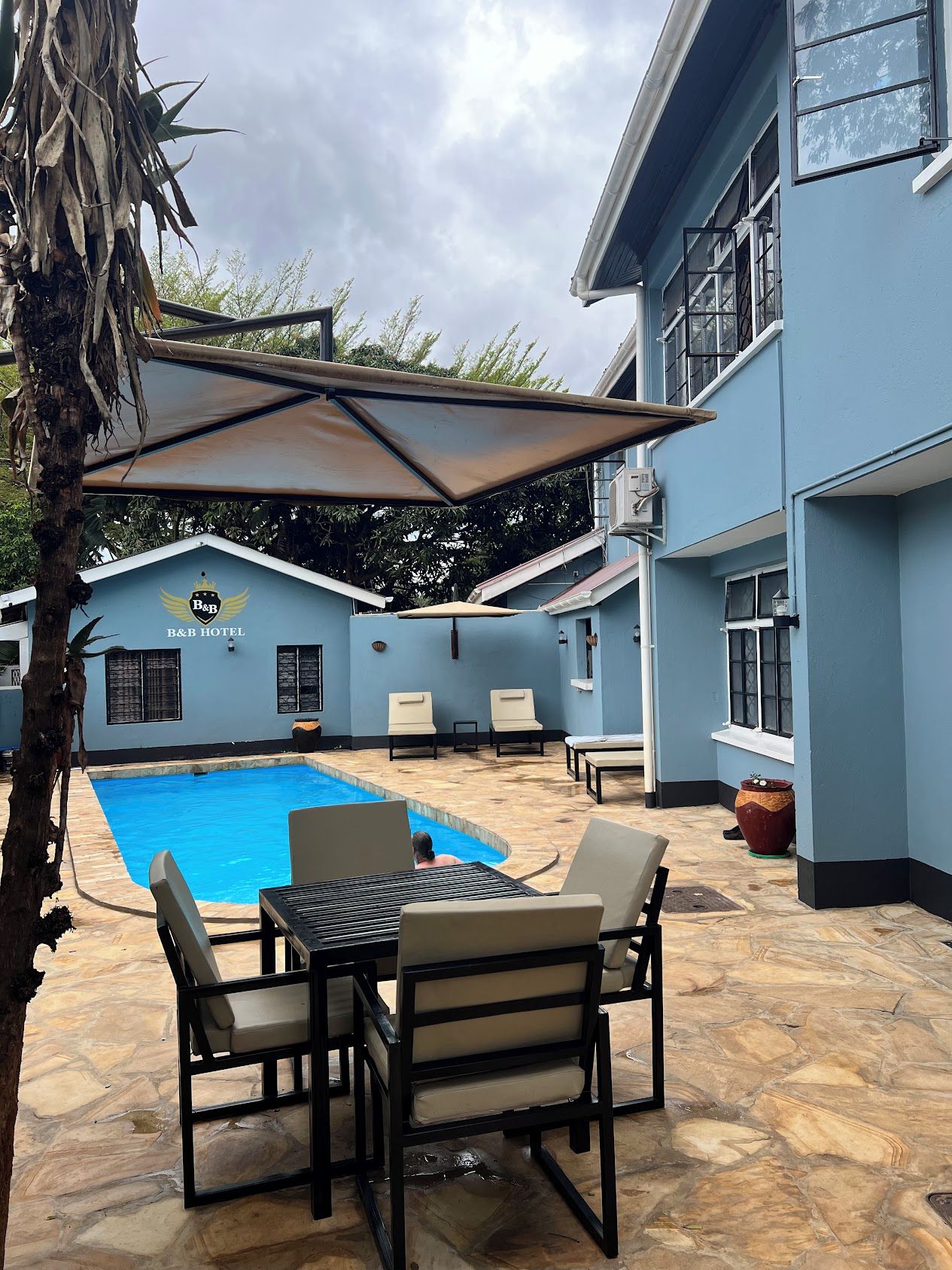 A pool area with lounge chairs and a table, a palm tree, and a building with a B&B logo.
