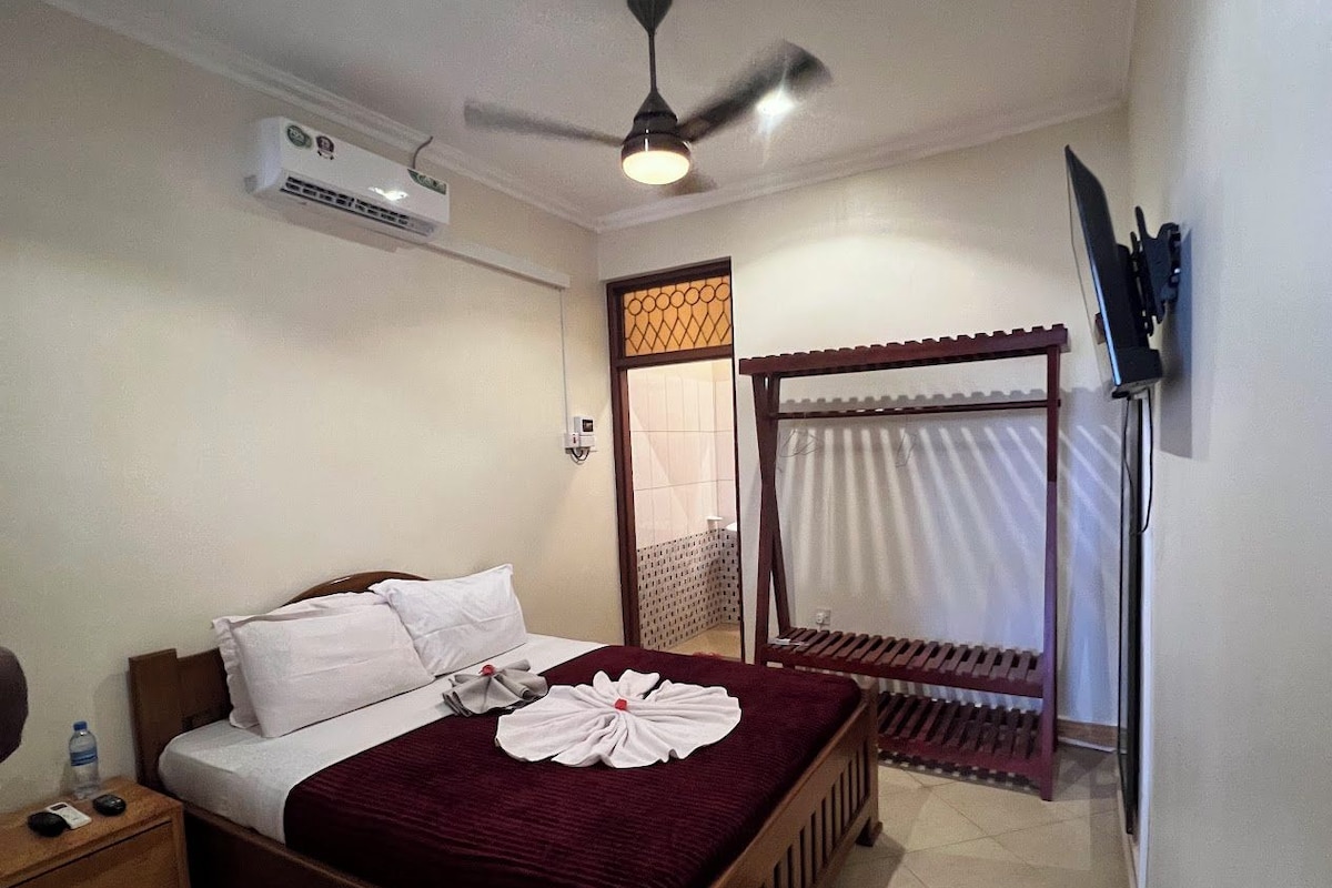 A hotel room with a bed, a ceiling fan, an air conditioning unit, a TV mounted on the wall, and a window with a curtain.