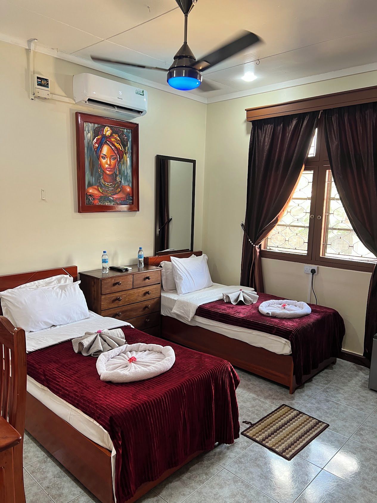 Habitación de hotel con dos camas, ventilador de techo, equipo de aire acondicionado, un cuadro, un espejo y una ventana con cortinas.