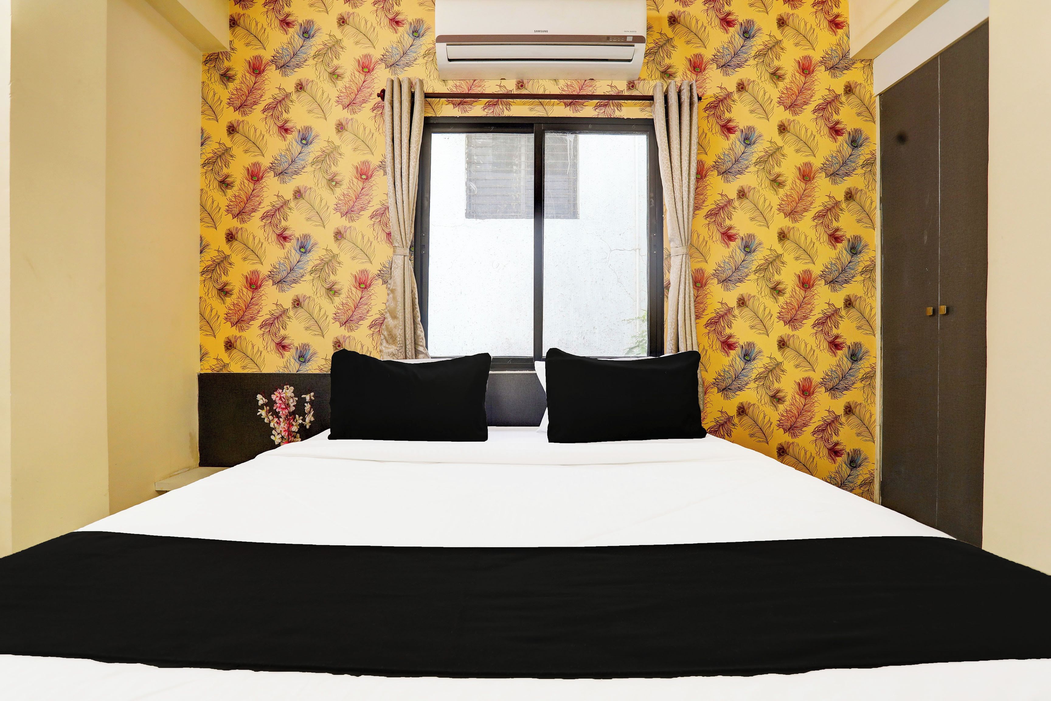 Habitación de hotel con cama, dos almohadas negras, ventana con cortinas y pared con estampado floral amarillo.