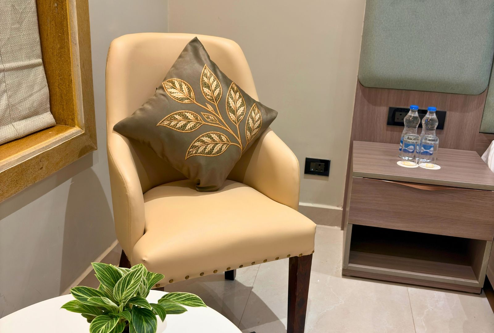 A modern armchair with a decorative pillow, a small table with a plant, and a wooden side table with water bottles and a plate.