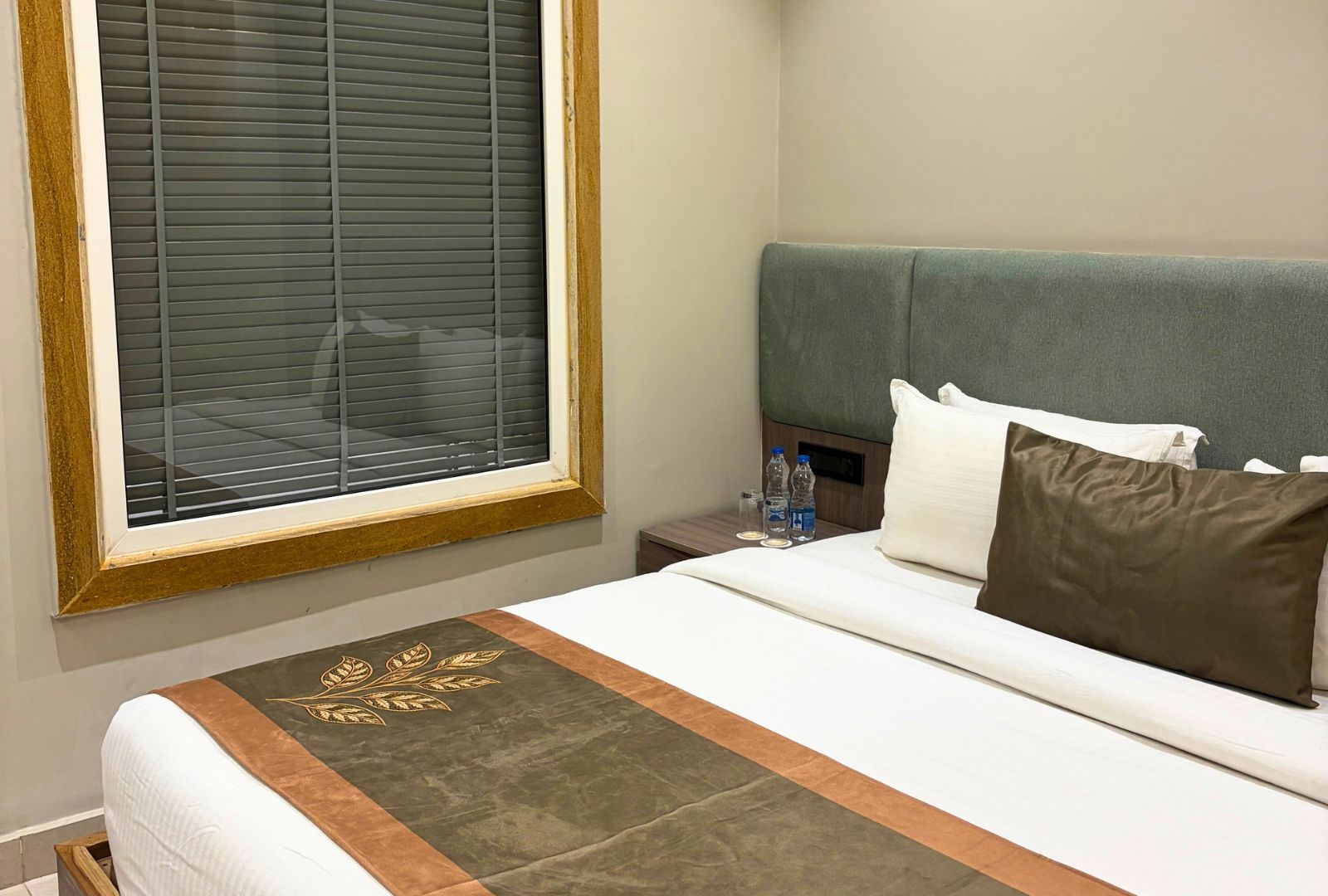 A hotel room with a bed, a headboard, a window with blinds, and a nightstand with water bottles.