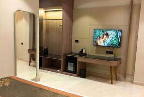 A hotel room with a large mirror, a flat-screen TV mounted on the wall, a small desk with a coffee maker, and a seating area with a bench. - Golden Bagh (Alwar)