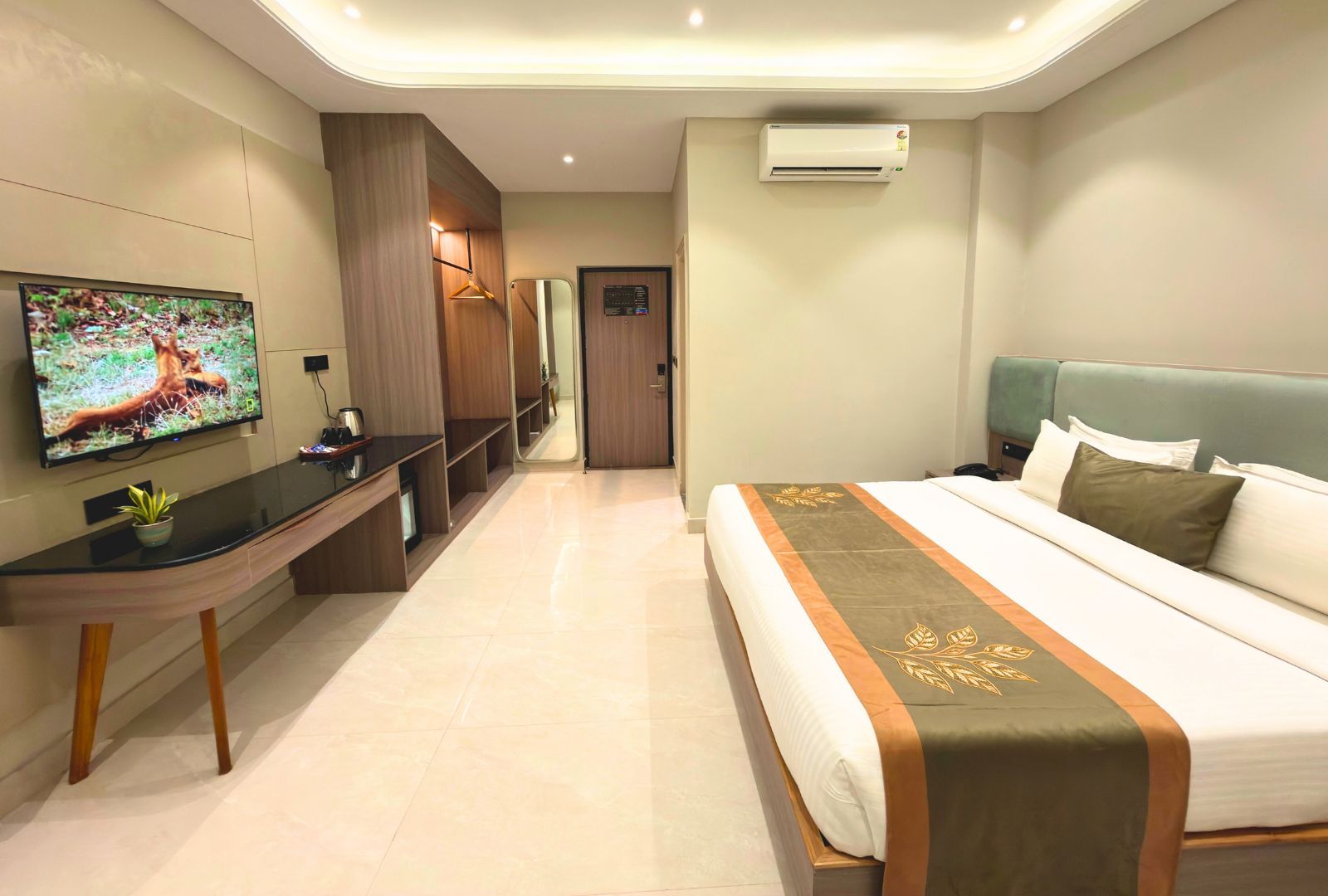 A hotel room with a bed, a desk with a TV, a mirror, and an air conditioning unit.