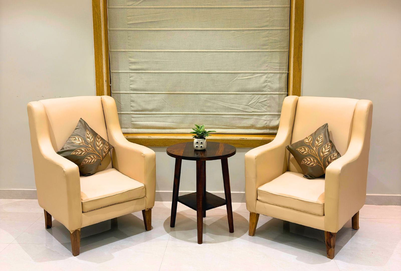 Two armchairs with decorative pillows, a small round table with a plant and a decorative object, and a large window with a blind.
