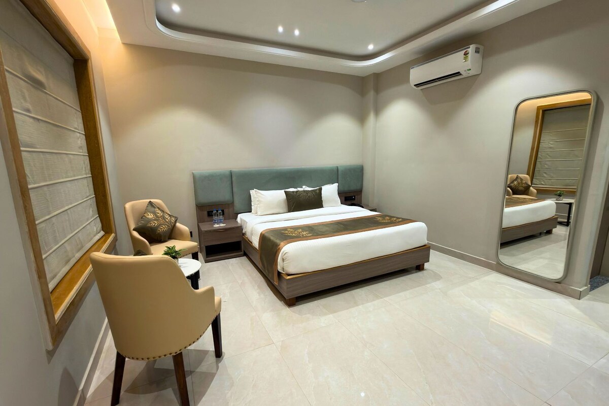 A modern hotel room with a large bed, a seating area with a chair and table, a bedside table with a lamp, and a large mirror.