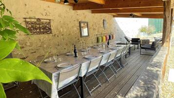 Une terrasse extérieure en bois, avec une longue table dressée pour les repas, entourée de chaises, et un mur de pierres rustiques en arrière-plan.