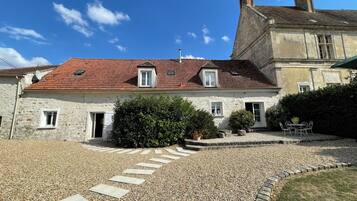 Un bâtiment en pierre avec un toit recouvert de tuiles rouges, une allée gravillonnée et un petit jardin agrémenté d’une table et de chaises.