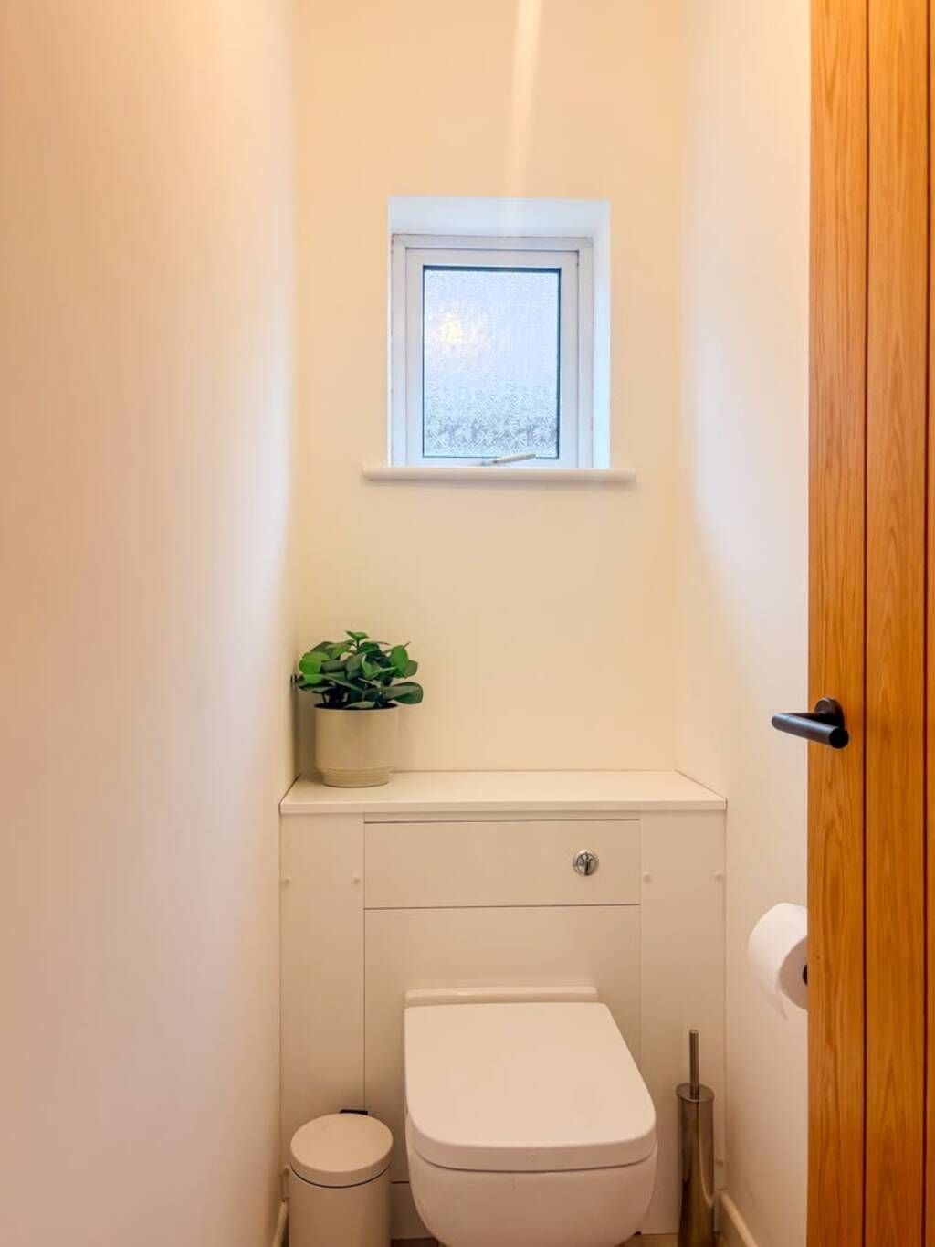 A small, clean bathroom with a toilet, a small trash bin, a window, and a potted plant on a cabinet.