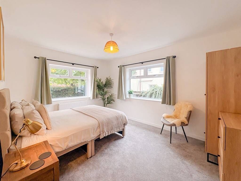 A bedroom with a bed, bedside table, lamp, chair, wardrobe, and windows with curtains.