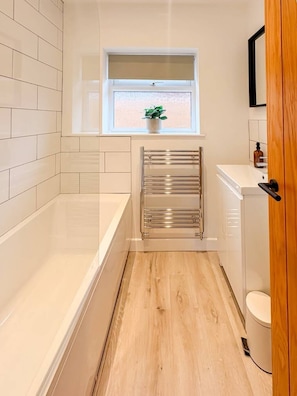 A narrow bathroom with a bathtub, a towel rack, a window with a plant, and a toilet. - Bungay Bungalow (Ditchingham)