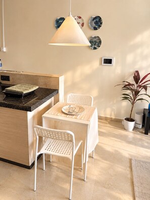 A modern kitchen with a central island, white chairs, a hanging light fixture, decorative plates on the wall, and a potted plant. - The Cozy House 1BHK Near BKC (Mumbai)