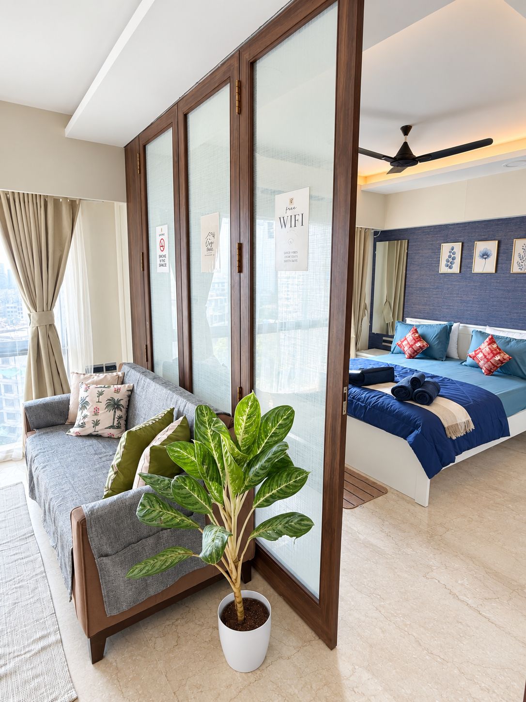Habitación de hotel con sofá, cama con ropa de cama azul, planta en maceta y puerta corredera con un cartel que indica WiFi gratuito.