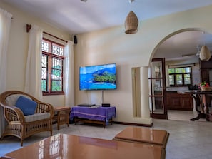A living room with a television, wicker furniture, a table, and a kitchen area visible through an archway. - 4-bedroom Beach Villa in superb Diani Beach with Private Pool & spectacular view (Diani Beach)