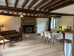 Un salon chaleureux avec une cheminée en pierre, des poutres apparentes, un canapé et un coin repas comprenant des chaises et une table.