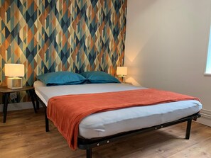 Ein Bett mit weißer Matratze und orangefarbener Decke, das von zwei Nachttischen mit Lampen gesäumt wird, in einem Raum mit geometrischem Wandpapier.