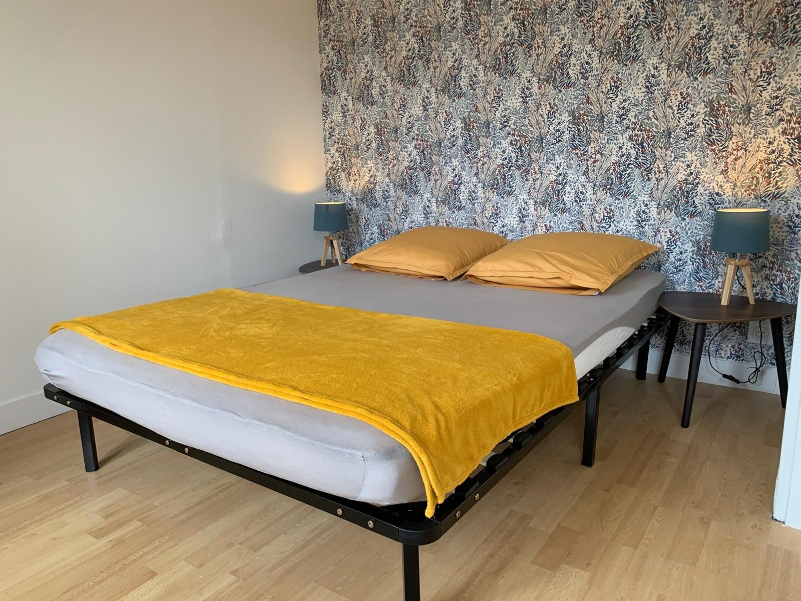Een bed met een gele deken, twee bedlampjes en behang met een patroon.
