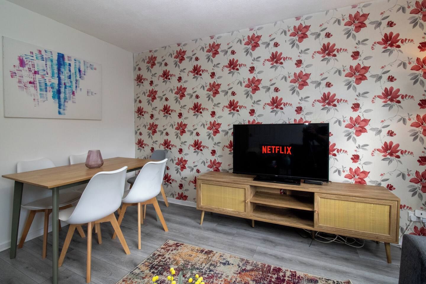 Un soggiorno con TV a schermo piatto che mostra 'NETFLIX', un tavolo da pranzo con sedie, carta da parati a motivi floreali e un tappeto.