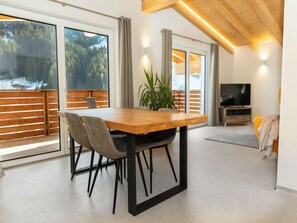 Un intérieur moderne avec une table à manger en bois, des chaises, une plante en pot et un téléviseur dans un espace de vie doté de grandes fenêtres et offrant une vue sur un paysage enneigé.
