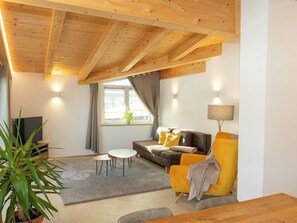 Un salon moderne avec un plafond en bois, un canapé, un fauteuil jaune, une petite table, un téléviseur et une fenêtre avec des rideaux.