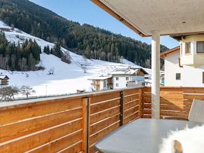 Een houten balkon met een tafel en stoelen, met uitzicht op een besneeerd landschap met huizen en een bebost gebergte.