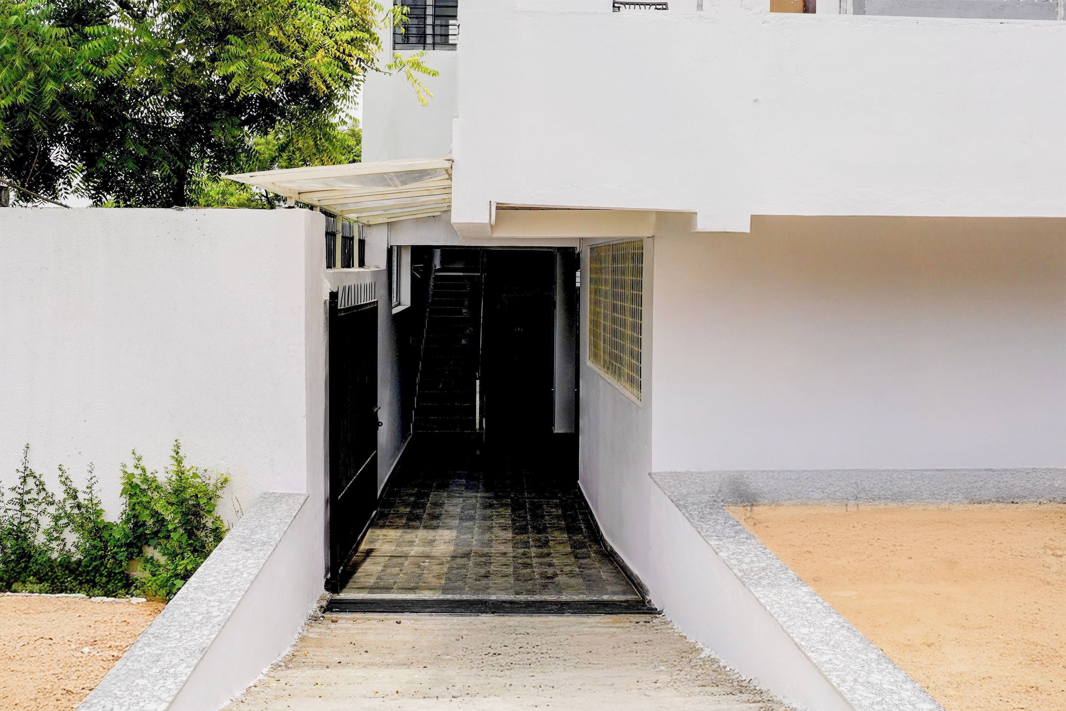 Ein schmaler, überdachter Weg führt zu einem Gebäudeeingang mit einem Metallgitter.