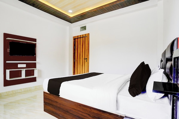 A hotel room with a bed, a TV mounted on the wall, a wooden door, and a ceiling with recessed lighting.