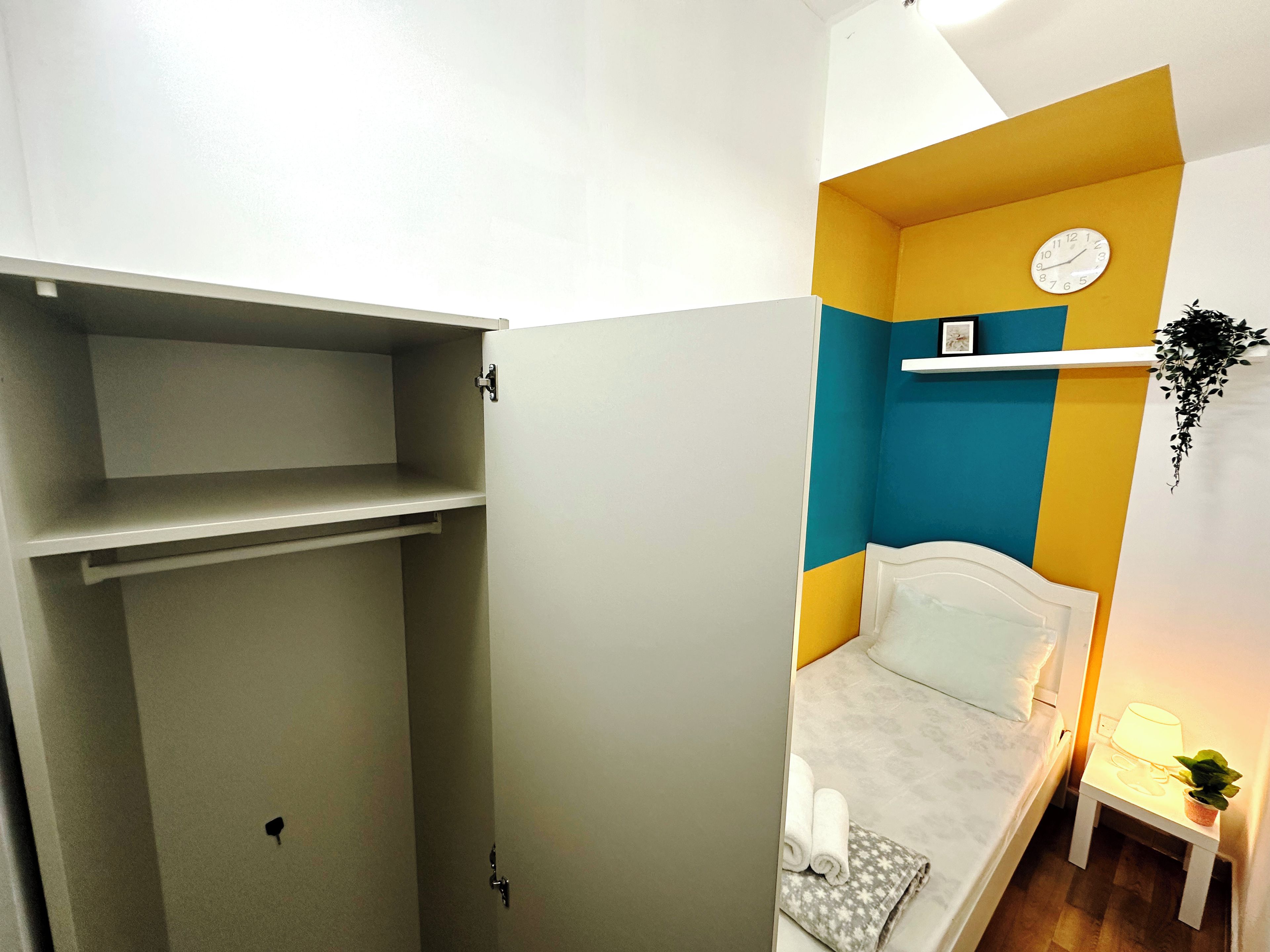 Una habitación con una cama, una mesita de noche con una lámpara, una pequeña planta en maceta y un reloj de pared.