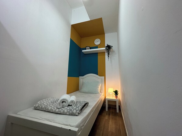 A small, narrow room with a bed, a bedside table with a lamp, and a wall clock.