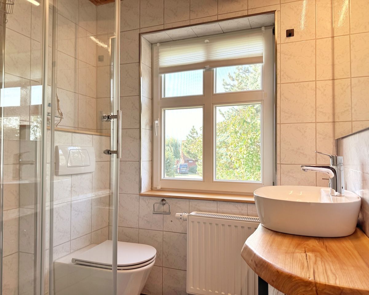Ein Badezimmer mit Duschkabine, Toilette, Waschbecken mit Holzablage und einem Fenster mit Jalousien.