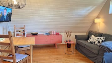 Ein gemütliches Wohnzimmer mit einem Holzesstisch, einer rosa Anrichte, einem grauen Sofa und einem an der Wand befestigten Fernseher.