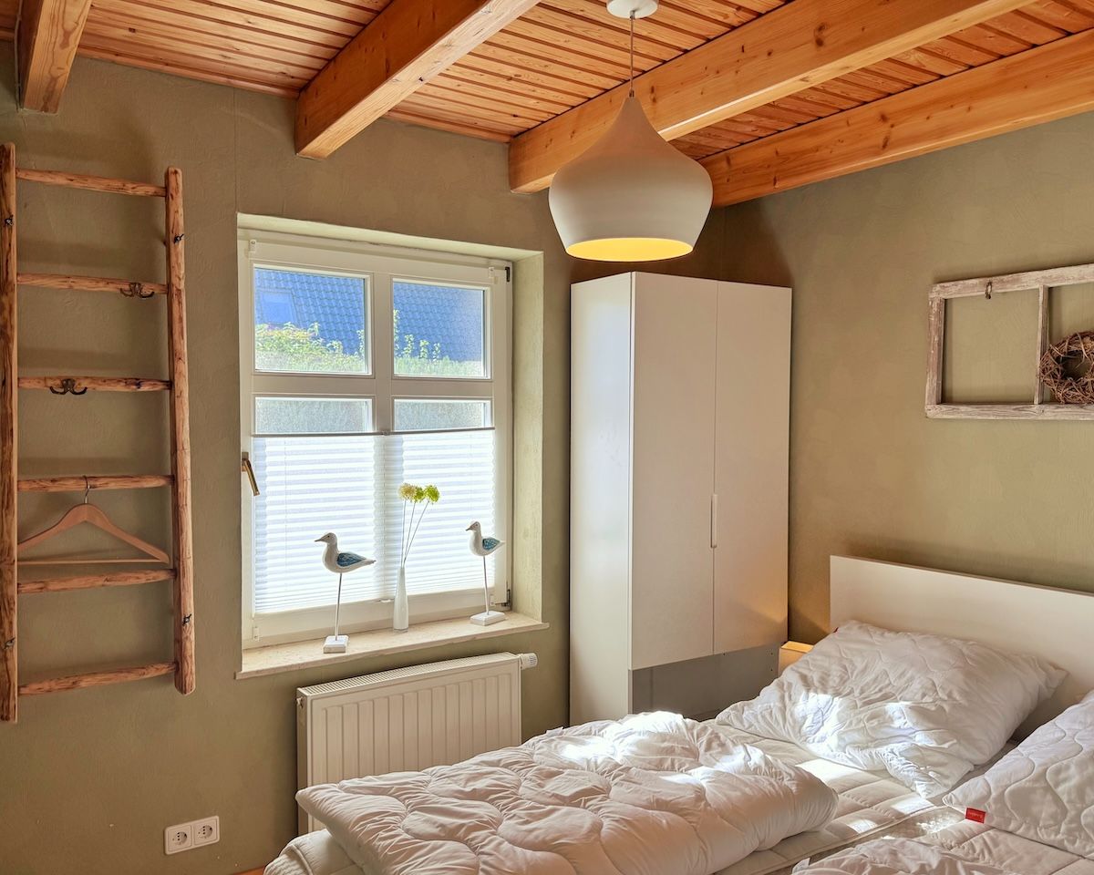 A bedroom with a wooden ceiling, a window with blinds, a white wardrobe, and a bed with white bedding.