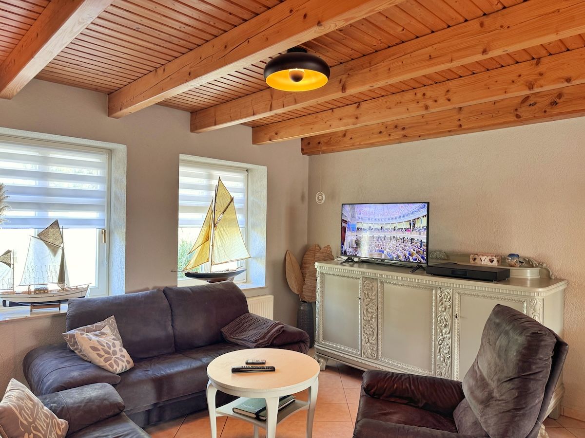 A living room with a brown leather sofa, a white coffee table, a television on a cabinet, and a sailboat model by the window.