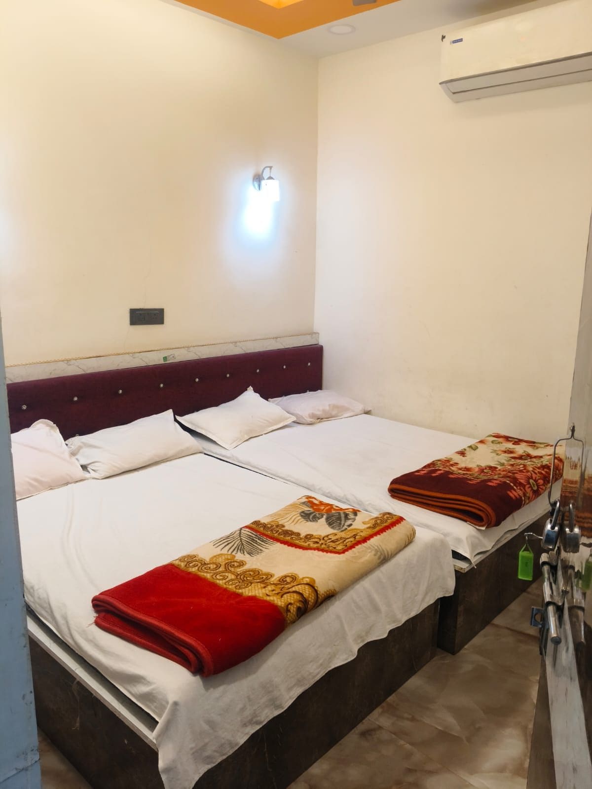 Ein Doppelbett mit weißen Leinentüchern und einer roten und goldenen Decke, ein Marmorkopfschild, eine an der Wand befestigte Lampe und eine Klimaanlage.