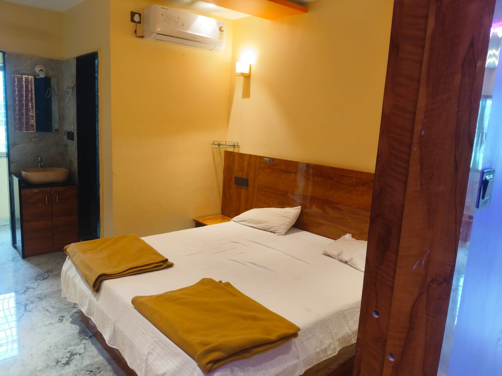 A hotel room with a bed, a wooden headboard, a nightstand, a wall-mounted air conditioner, and a bathroom with a sink and mirror.