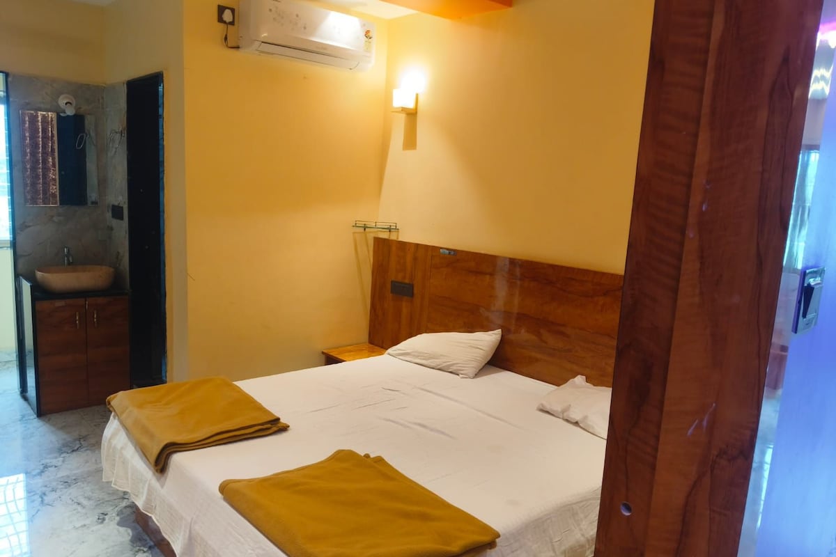 Habitación de hotel con cama, cabecera de madera, mesita de noche, aire acondicionado de pared y baño con lavabo y espejo.