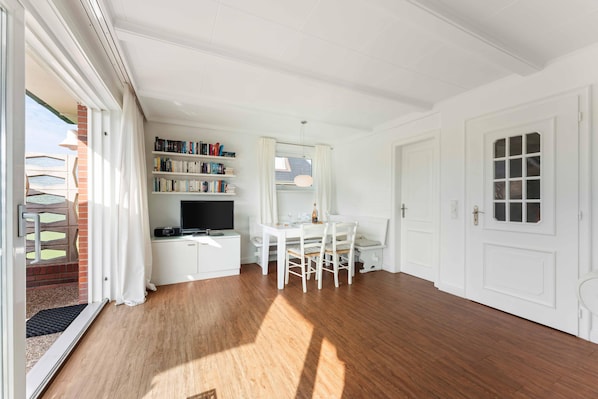 Una habitació lluminosa amb terra de fusta, una taula de dinar blanca amb cadires, una biblioteca i una televisió.