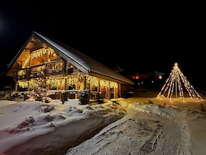 Une cabane en rondins dont le toit est recouvert de neige, avec un sapin de Noël illuminé au premier plan.