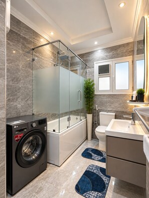 A modern bathroom with a glass-enclosed shower, a washing machine, a bathtub, and a sink. - Luxury Family 3BR Apartment - Steps to Pyramids, Grand Egyptian Museum & Saqqara (Giza Governorate)