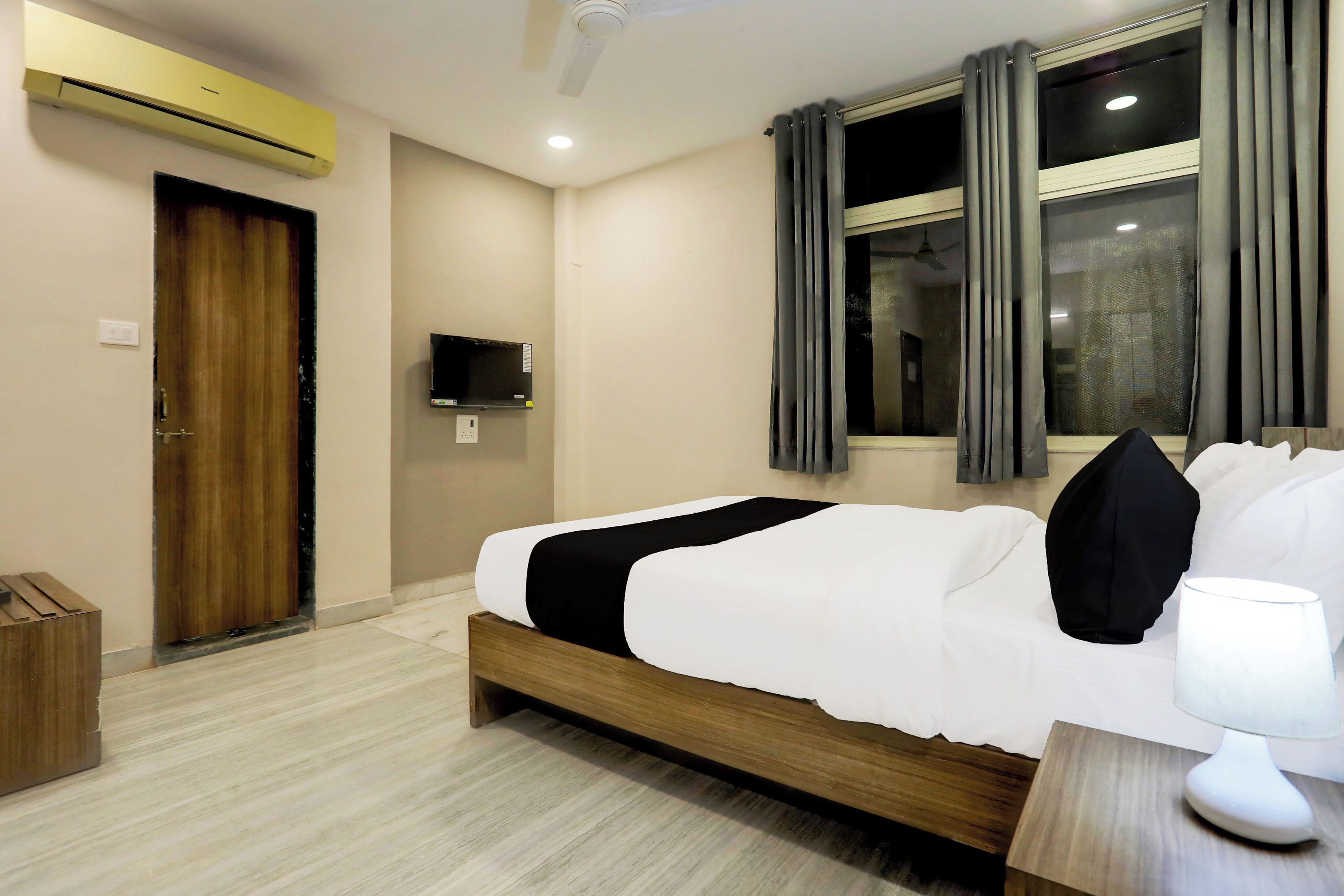 A hotel room with a bed, a bedside table, a lamp, a window with curtains, an air conditioning unit, a television, and a door.