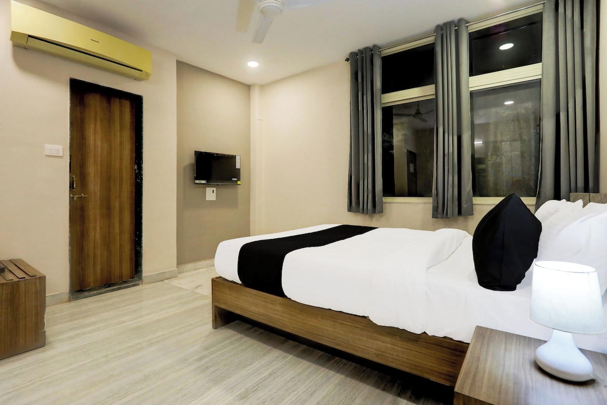 Habitación de hotel con cama, mesita de noche, lámpara, ventana con cortinas, equipo de aire acondicionado, televisor y puerta.