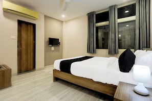 Habitación de hotel con cama, mesita de noche, lámpara, ventana con cortinas, equipo de aire acondicionado, televisor y puerta.