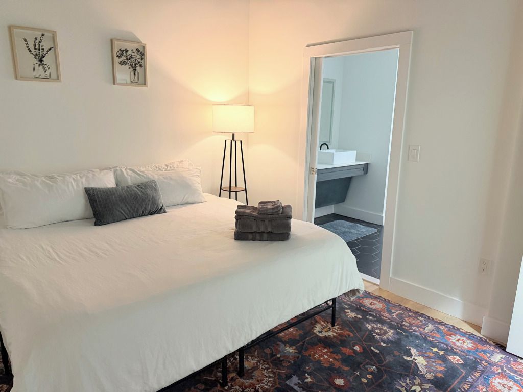 Un dormitorio con cama, una lámpara de pie, un tapete y un baño visible a través de una puerta abierta.